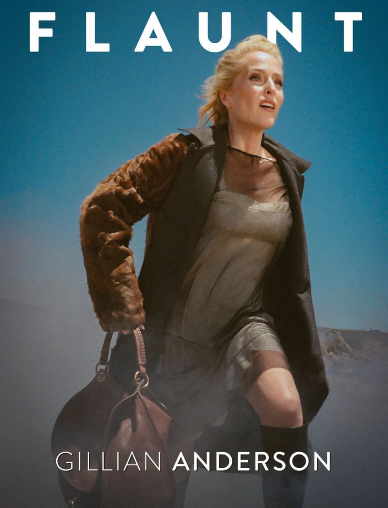Gillian Anderson walking forward against a clear blue sky, wearing a dark coat and sheer dress while carrying a brown leather handbag.