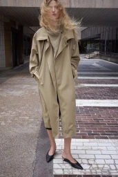 Model Felice Nova Noordhoff wearing an oversized beige trench coat and black pointed mules on an urban tiled path.