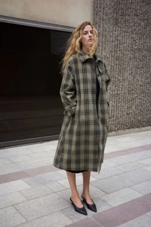 Model Felice Nova Noordhoff stands outdoors wearing a long olive green plaid trench coat and black pointed kitten heels.