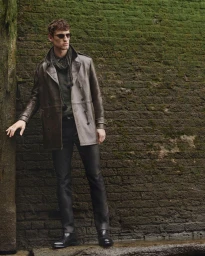 Model Anatol Modzelewski in a brown leather pea coat, dark trousers, and sunglasses standing against a mossy brick wall.