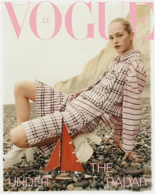 Vogue cover featuring a woman in pink plaid jacket and shorts, white platform boots, seated near a small sailboat on a pebble beach.