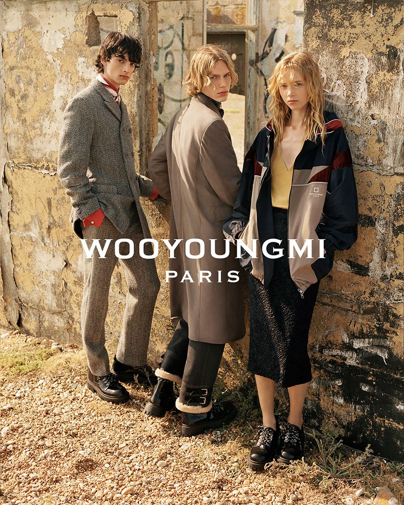 Three models in tailored outerwear, tweed suit, and an oversized zip-up jacket posing outdoors against a distressed concrete wall.