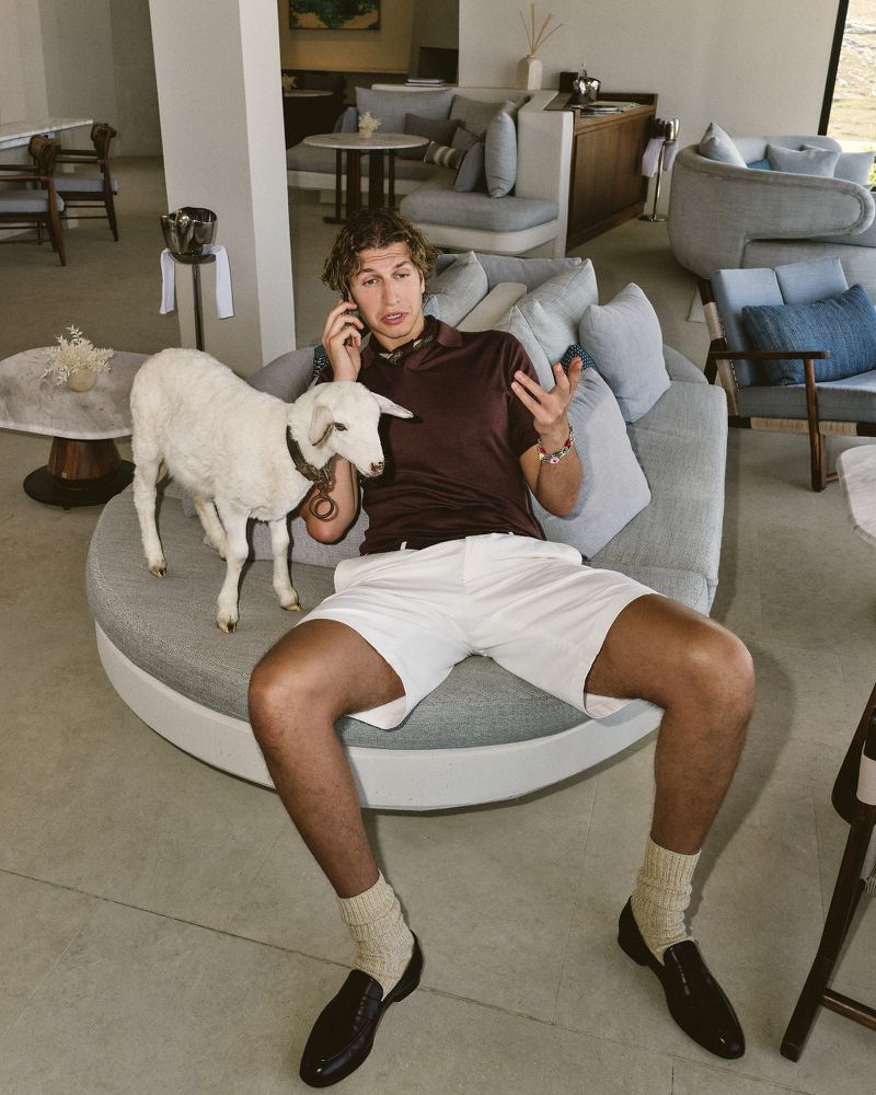 A man sits on a round sofa indoors, talking on a cell phone, while a white goat stands beside him on the sofa, bathed in natural light.