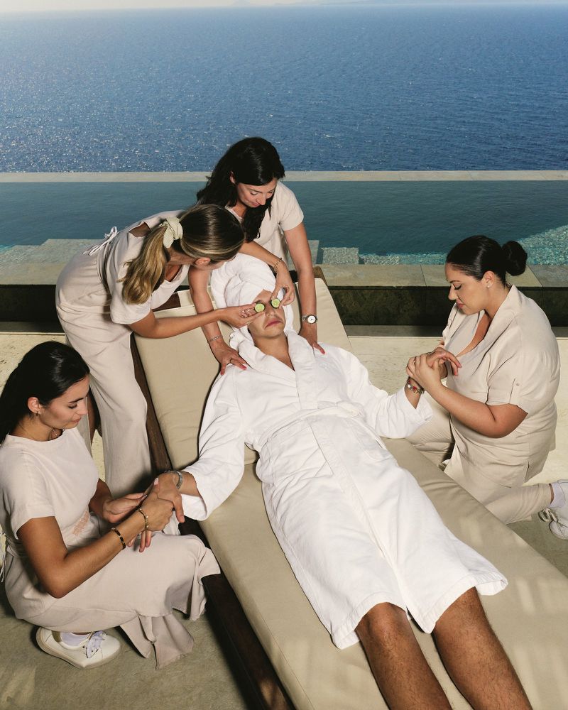 Person receiving spa treatment with cucumber slices on eyes and hands being massaged by several spa workers near infinity pool.