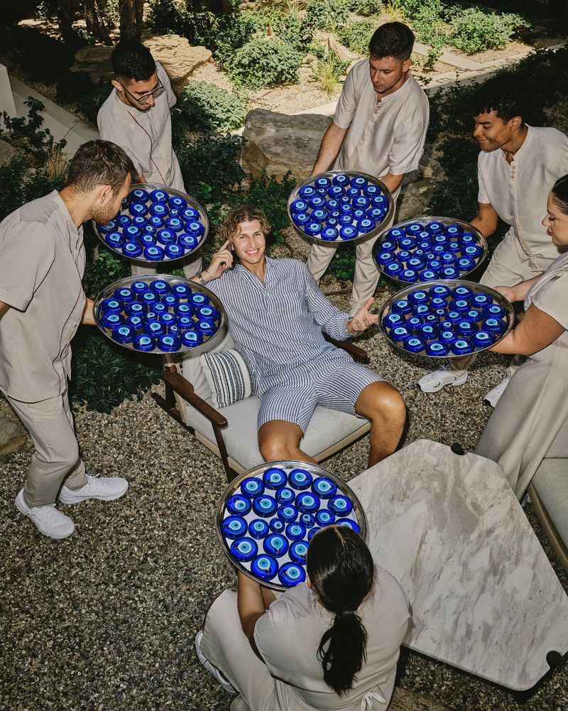 Man sits in a chair surrounded by servers holding trays filled with blue amulets, outdoors in a sunny, landscaped garden setting.