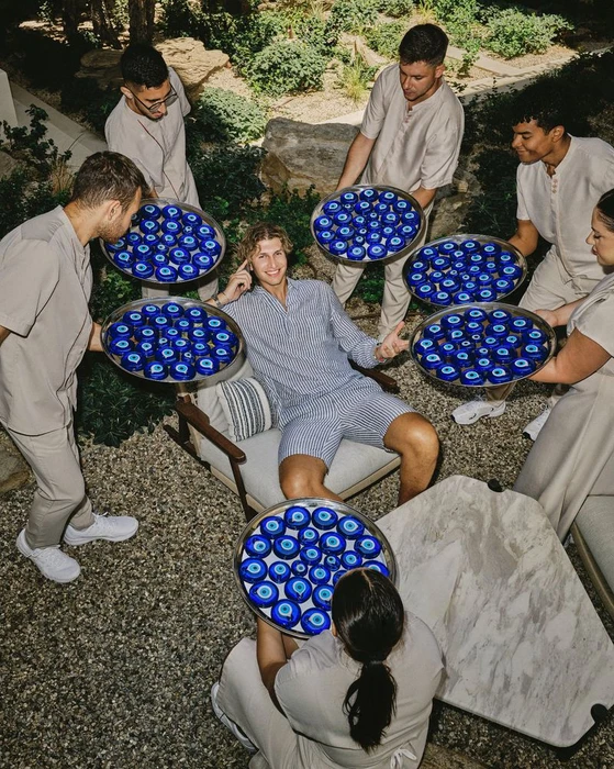 Man seated in an armchair surrounded by staff holding silver trays filled with blue evil eye amulets for Gstaad Guy and One&Only