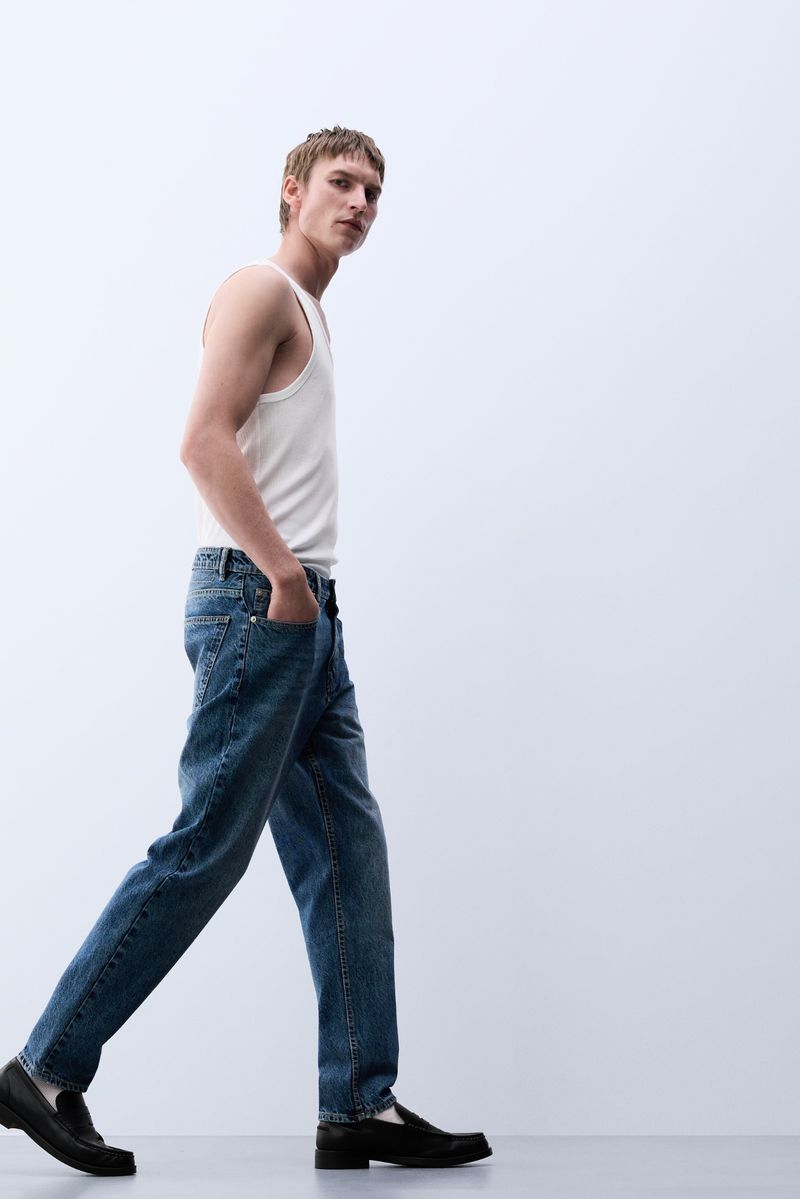 A young adult male model in jeans and a white tank top walks against a white backdrop, showcasing fashion and casual style.