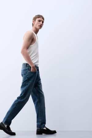 A young adult male model in jeans and a white tank top walks against a white backdrop, showcasing fashion and casual style.