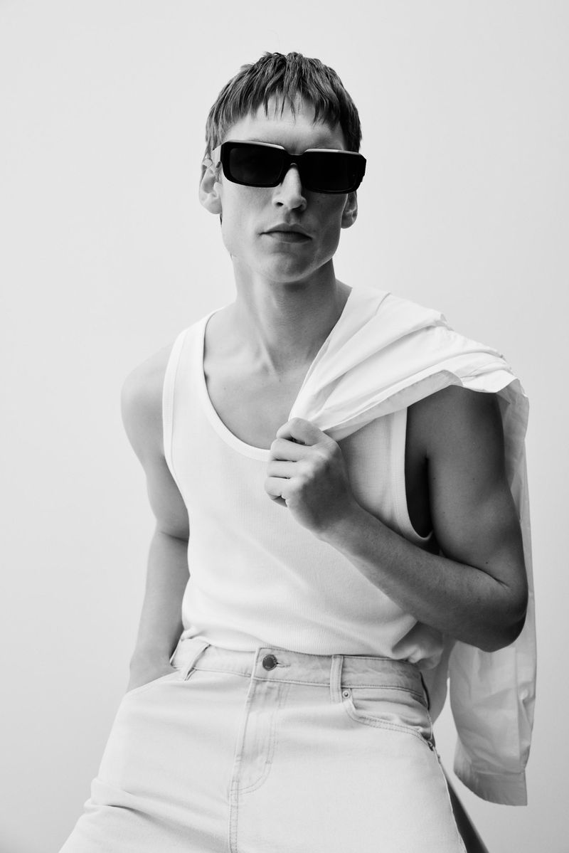 Black and white portrait of a young adult man wearing sunglasses, a white tank top, and light jeans. He has a white shirt slung over his shoulder.