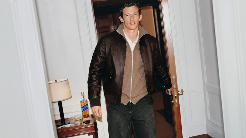 Man wearing a brown leather jacket and beige knit zip-up cardigan walks through a doorway beside a table with a bottle of Johnnie Walker.