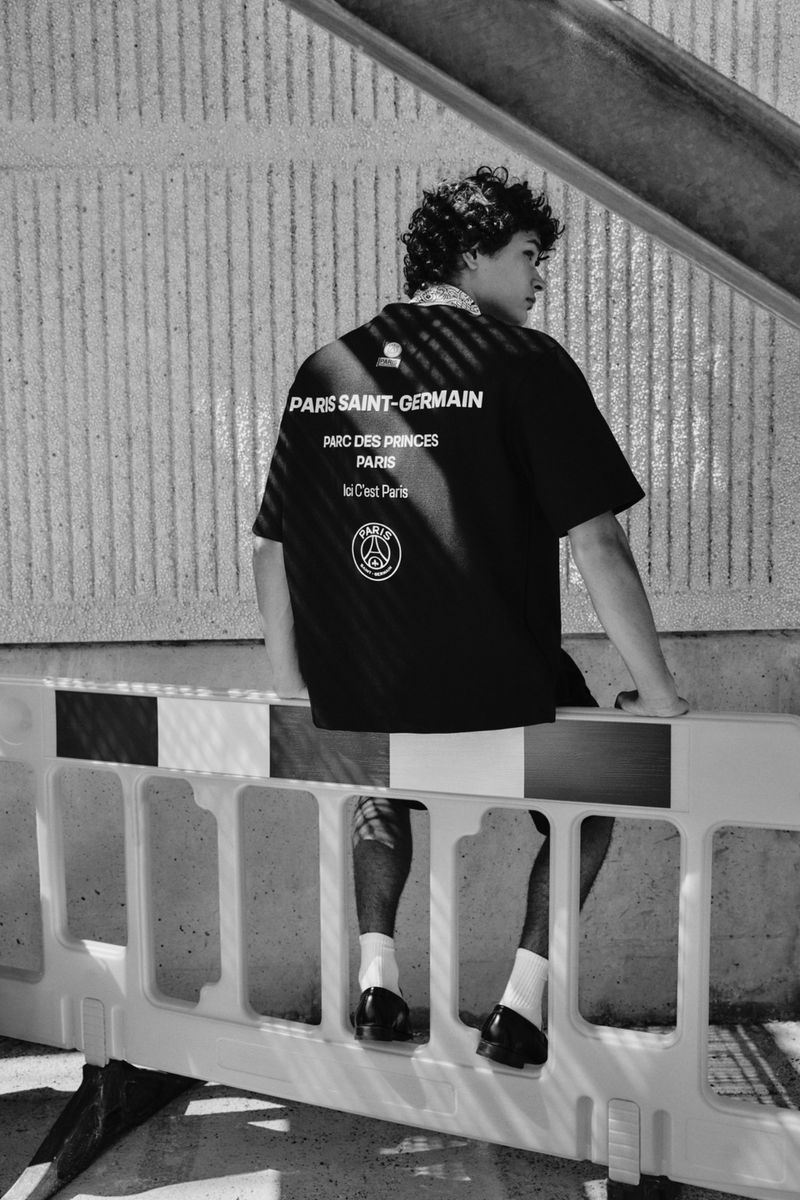 Black and white photo of a person in a black t-shirt with 'Paris Saint-Germain' text, sitting on a white and black plastic barricade outdoors.