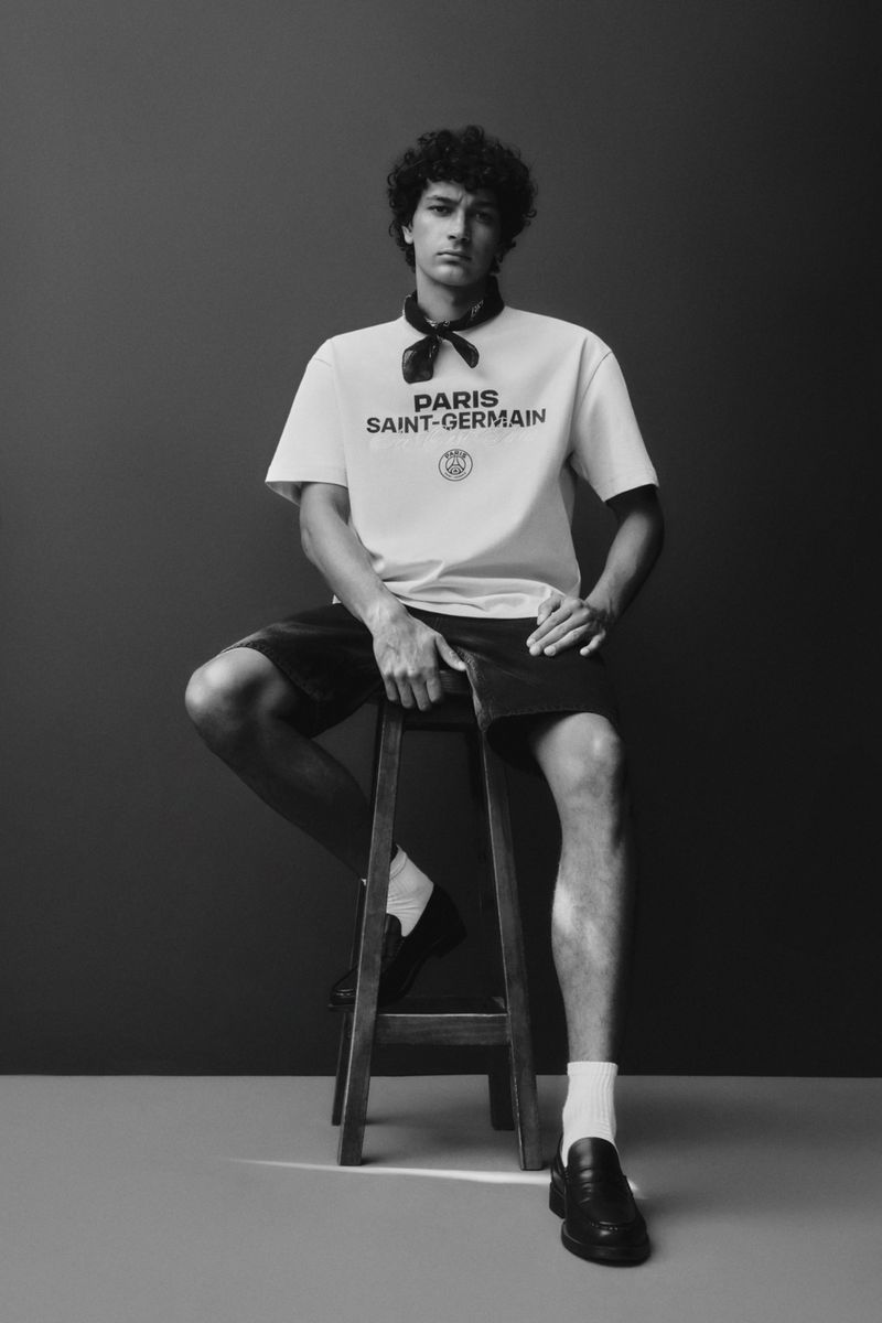 A young man sits on a wooden stool in a photo studio wearing a white T-shirt, dark shorts, loafers and a neck scarf in monochrome.