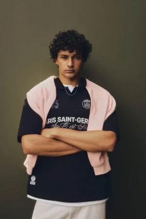 Young adult man in navy Paris Saint-Germain t-shirt and white pants, casually posing indoors with arms crossed against a neutral background.