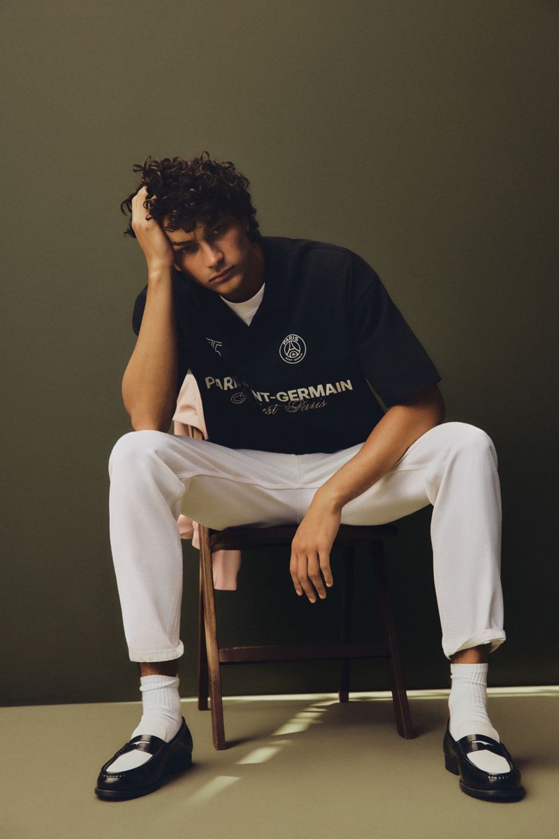A young adult male model with curly hair poses indoors while sitting on a wooden chair, wearing a black t-shirt, white sweatpants, and black loafers.