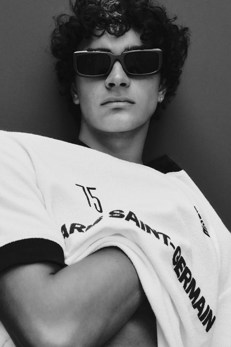 Portrait of a young adult male with curly hair, wearing sunglasses and a branded shirt, posing indoors against a plain dark background.