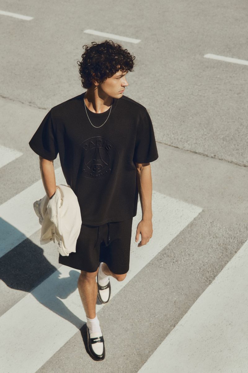 A young adult male walks in black shorts and matching t-shirt on a crosswalk with a jacket slung over his arm in daylight.