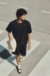Young man with dark curly hair wearing black t-shirt and shorts walking across a street with white road markings