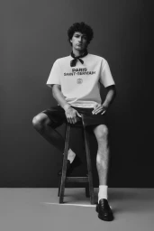 Model seated on a wooden stool wearing a branded t-shirt and dark shorts against a seamless grey backdrop