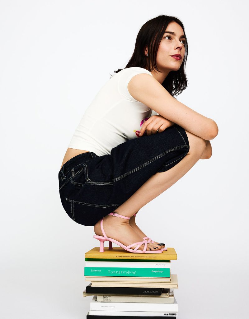 Brunette woman in white t-shirt and dark denim shorts crouching on stack of books, wearing pink high-heeled strappy sandals.