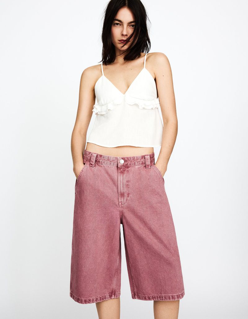 Brunette fashion model poses indoors wearing a white ruffle top and pink denim culottes against a neutral backdrop.