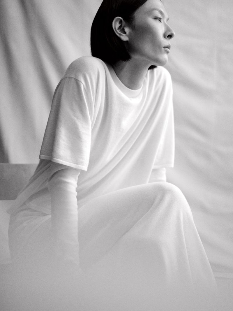 Model Hsin-Yu Liu seated in profile wearing layered white knitwear against a draped fabric backdrop in a black and white portrait.