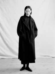 Full-length black and white portrait of a woman in an oversized black tailored overcoat standing against a draped fabric backdrop.
