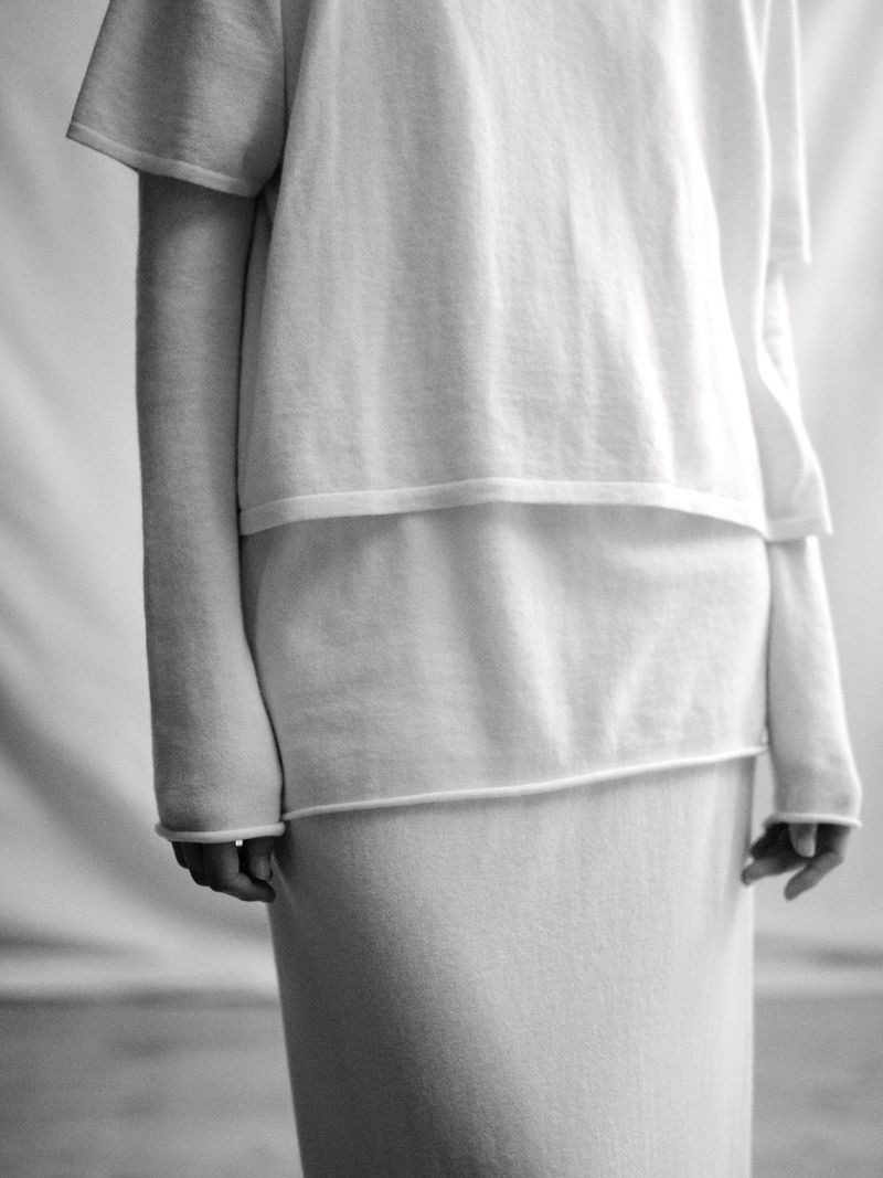 Close-up of a model wearing layered white knit tops and a matching skirt against a draped fabric studio backdrop.