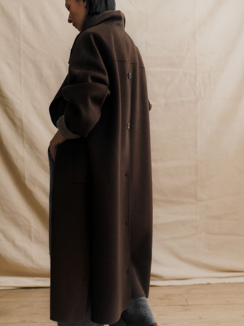 Model Hsin-Yu Liu wearing an oversized long brown coat with back buttons against a warm canvas backdrop.