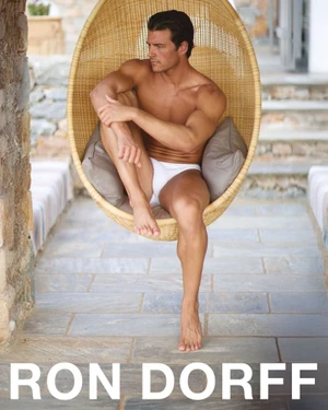 Model Aiden Stonehouse seated in a hanging woven chair wearing white swim briefs on an outdoor stone patio.