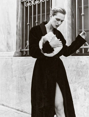 Model Nikki McGuire in a dark textured coat holding a white quilted clutch bag against an exterior stone wall.
