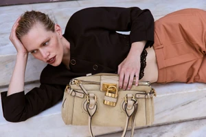 Model Nikki McGuire reclining on marble steps in a dark cropped blazer and terracotta pants holding a beige padlock handbag.