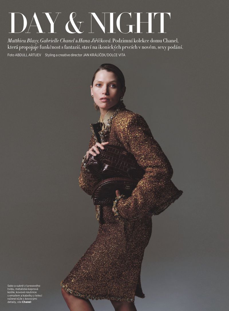 Model Hana Jirickova posing in a shimmering bronze tweed skirt suit holding a textured clutch against a grey studio backdrop.