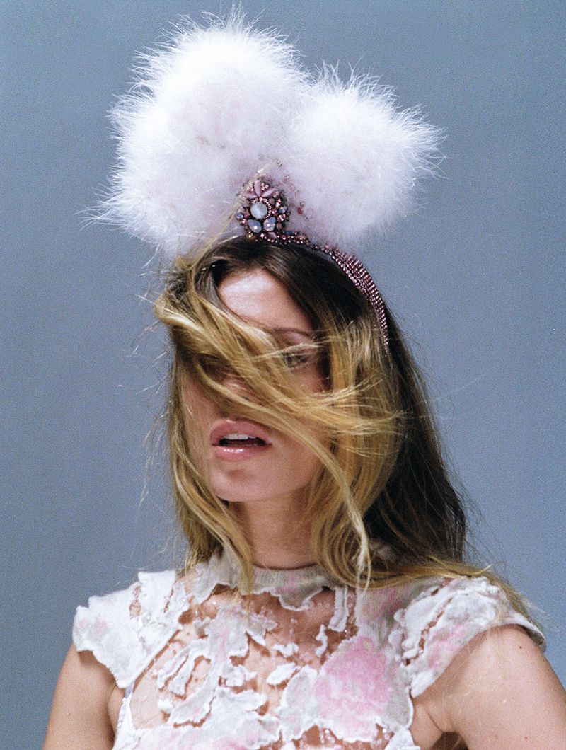 A woman wears a headband topped with two large, fluffy white feather pom-poms and a crystal-encrusted, pink-toned ornate crown base.
