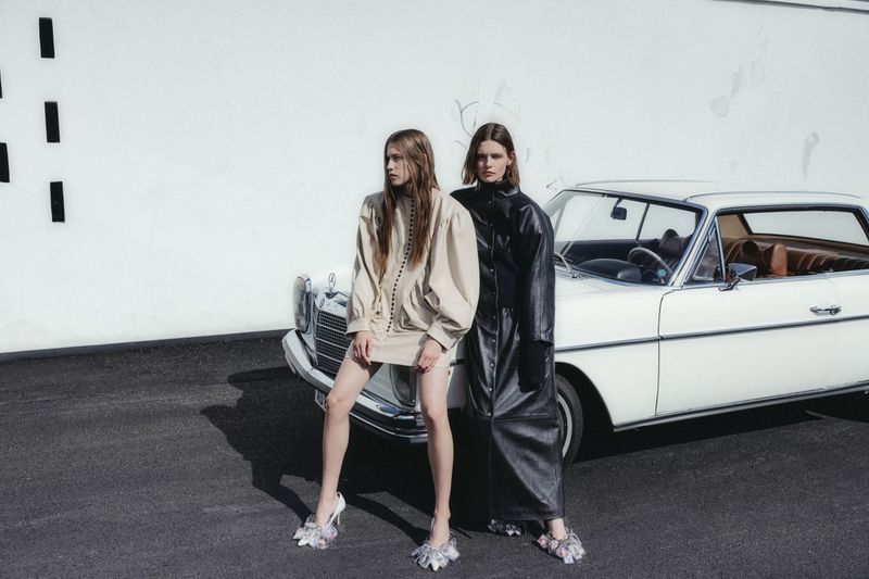 Two models in a beige puff-sleeve dress and a long black leather coat pose beside a vintage white car against an exterior white wall.
