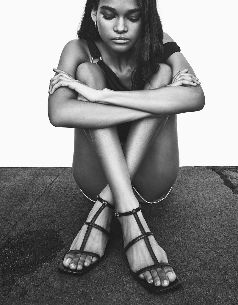 Portrait of an adult female sitting, embracing her legs, and wearing strappy high-heel sandals in a minimalist black and white photo.
