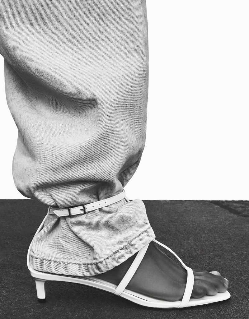 Close-up studio shot of a foot wearing a white heeled sandal with strappy design. The ankle is encircled by a thin white belt.