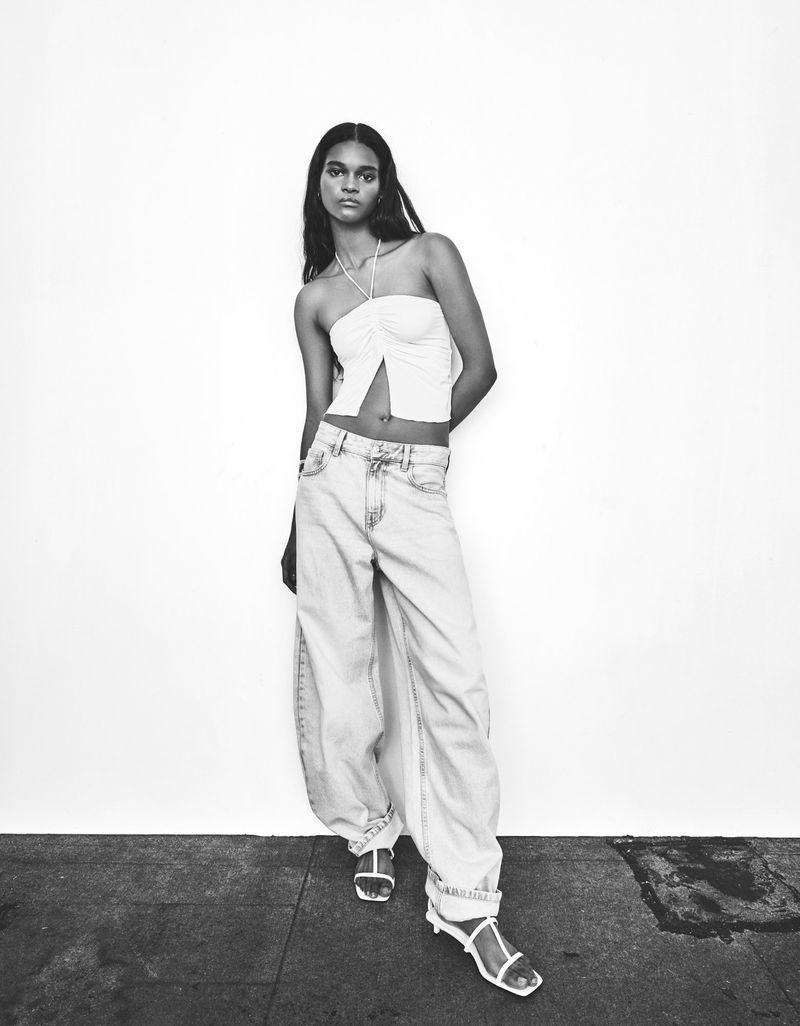 Black and white portrait of a young woman in a halter top and light-wash jeans posing against a neutral wall in a full-body shot.