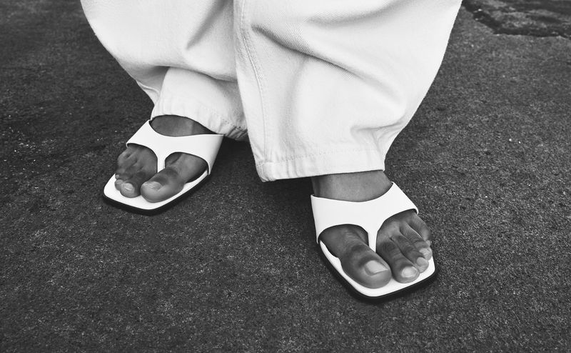 Feet wearing white thong sandals and white pants stand on a concrete surface in a black and white photograph; fashion footwear.