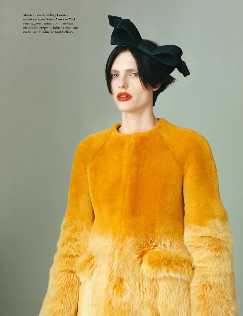 Model wearing a yellow ombre textured fur coat and an oversized dark bow headpiece against a pale neutral background.
