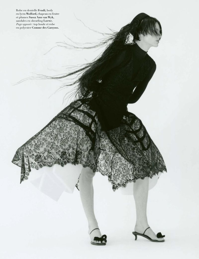 Model Charlie Jones poses dynamically in a black lace structured skirt and feathered headpiece against a white background.