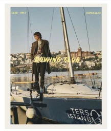 Model Sasha Cortesi standing on a sailboat deck against a sunlit coastal city skyline in an editorial portrait.