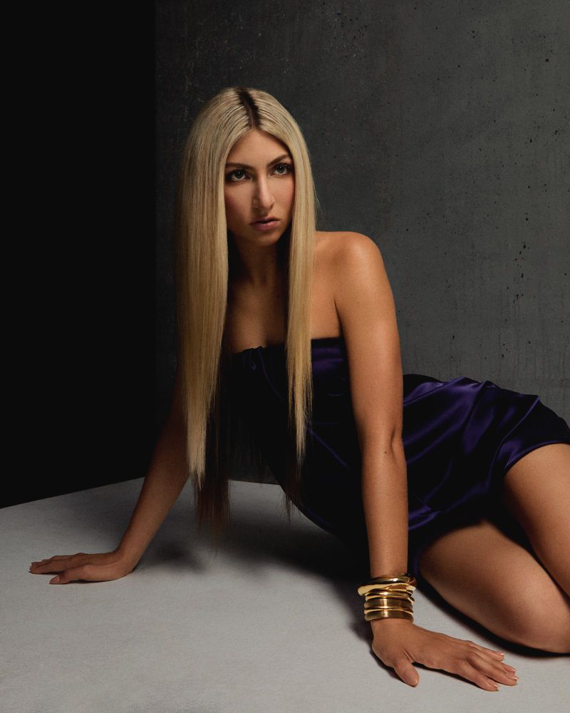 Blu DeTiger with long blonde hair in a purple satin strapless mini dress, wearing gold bangles, posed on a concrete studio floor.