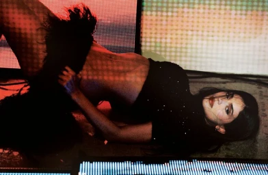 Kylie Jenner lying on the floor surrounded by glowing digital LED screens in a black cropped top and textured feather skirt.