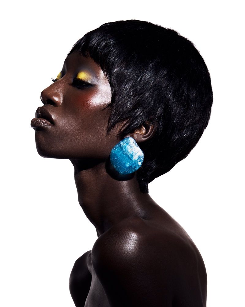 Profile portrait of a Black woman with short black hair, yellow eye makeup, and a blue statement earring against a white background.