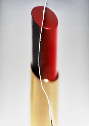 Close-up of a red lipstick in a gold tube vertically divided by a white thread against a neutral backdrop.