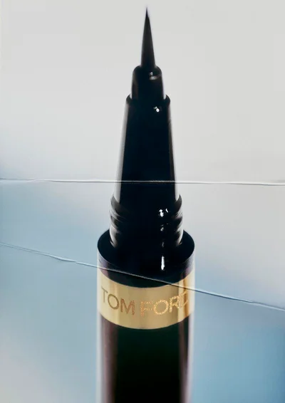 Close-up of a Tom Ford liquid eyeliner pen with a fine pointed tip and gold logo band behind glass on a pale blue background.