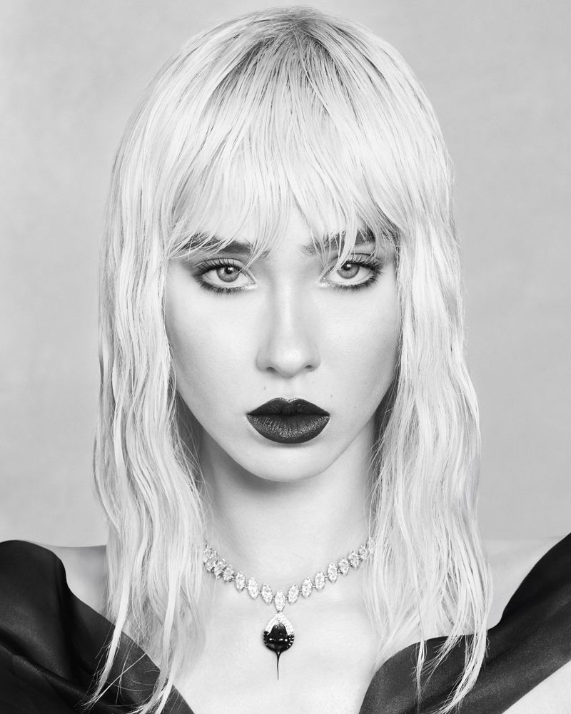 Black and white portrait of a woman with textured blonde bangs wearing a sparkling pendant necklace and dark off-the-shoulder attire.