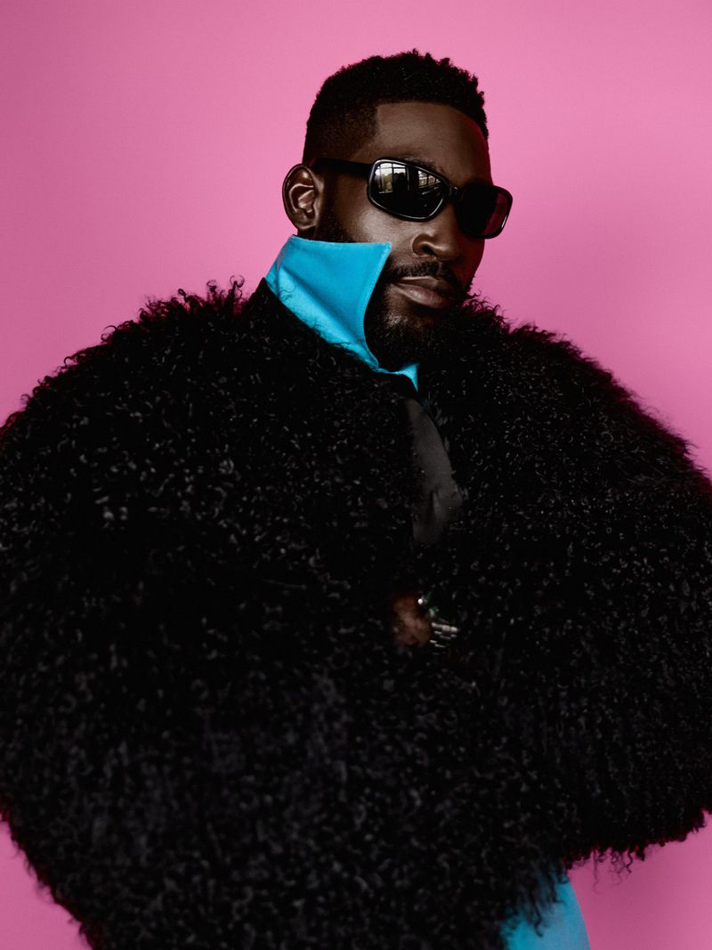 Musician Tinie Tempah wearing a voluminous black textured coat, blue high collar, and dark sunglasses against a bright pink backdrop.