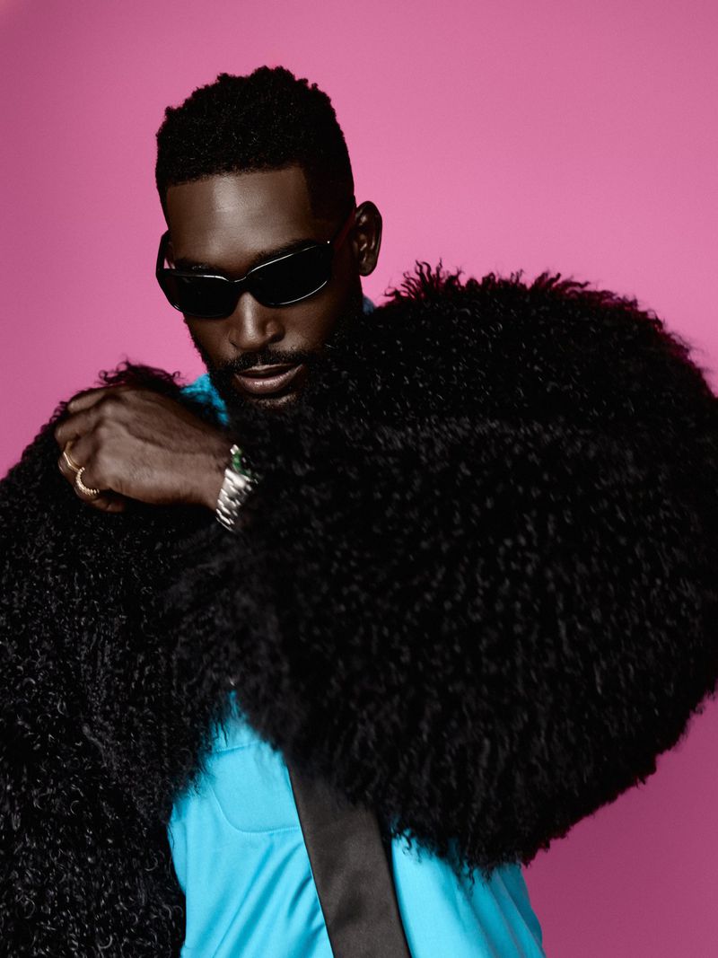Tinie Tempah wearing dark sunglasses and an oversized black shearling coat against a pink background.