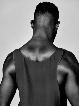 Monochrome studio portrait of Tinie Tempah seen from behind wearing a dark sleeveless tank top against a plain studio backdrop.